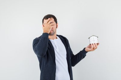 young man in white t-shirt and zip-front black hoodie holding house model and covering face with hand and looking harried , front view.